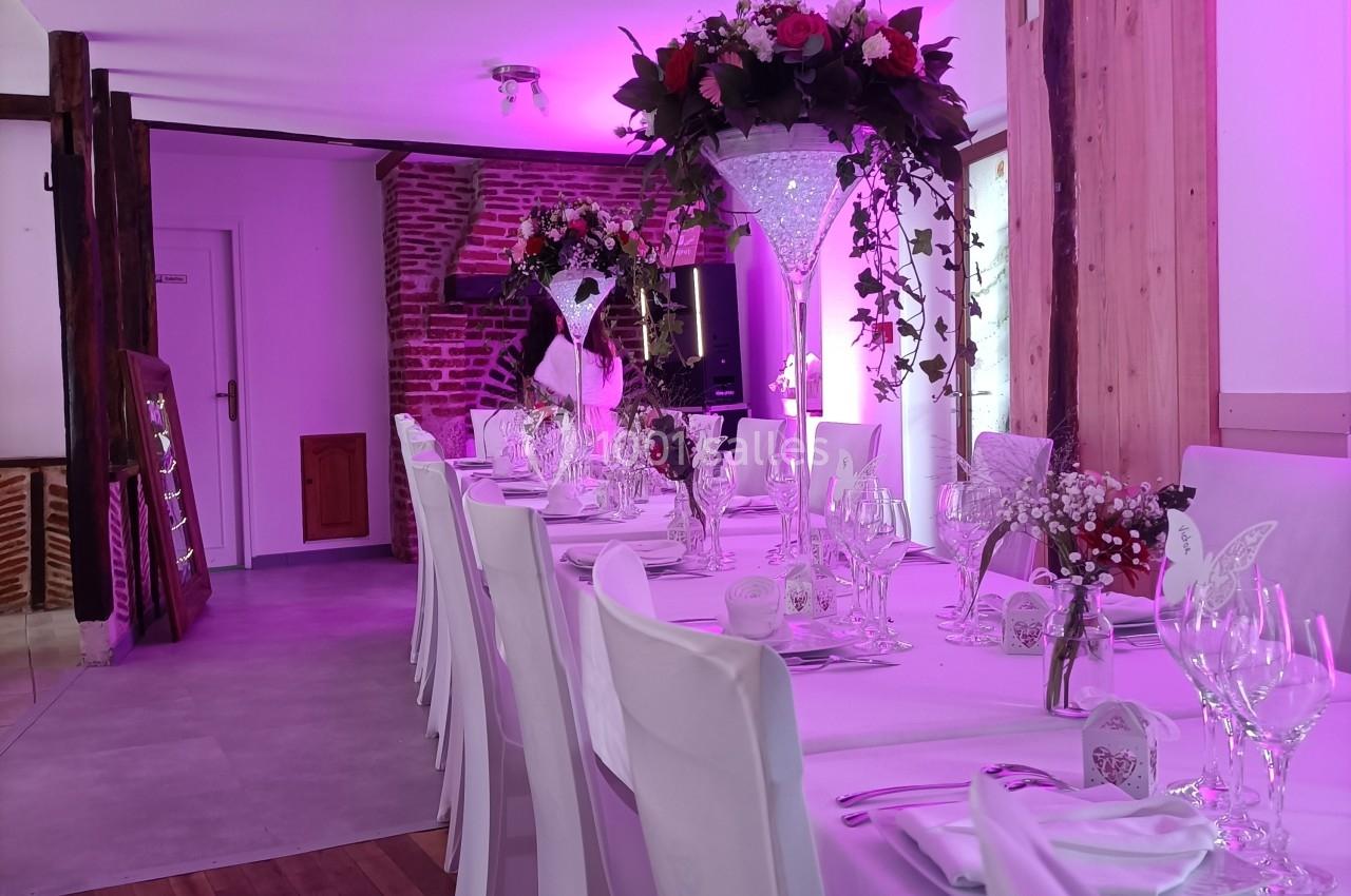 Salle de réception décorée avec une longue table dressée, nappes blanches, fleurs et éclairage rose tamisé.