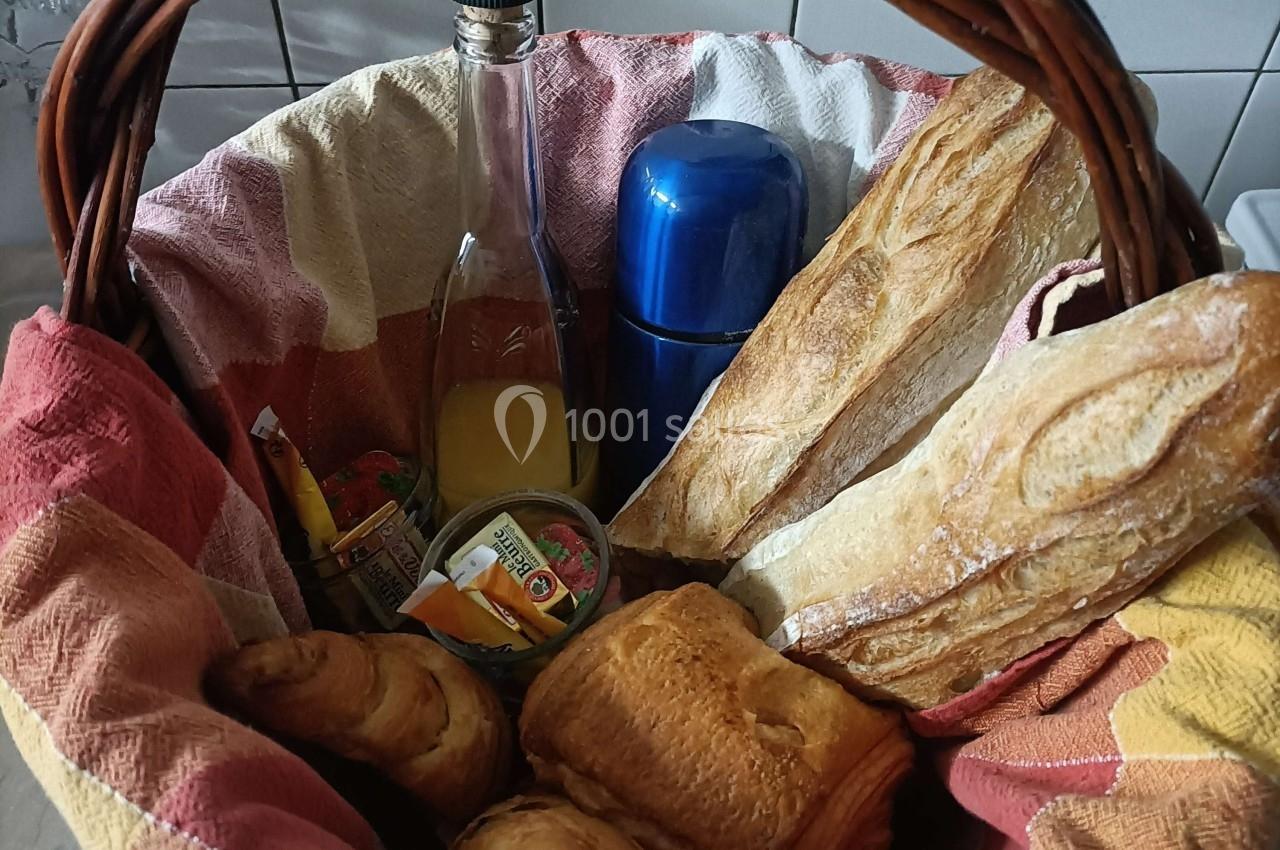 Panier en osier contenant des baguettes, viennoiseries, jus de fruit, thermos et confitures, posé sur une table.