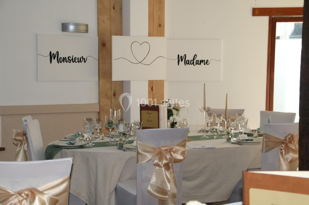 Table de réception décorée avec nappes claires, chaises ornées de nœuds dorés et panneaux ’Monsieur’ et ’Madame’ au mur.