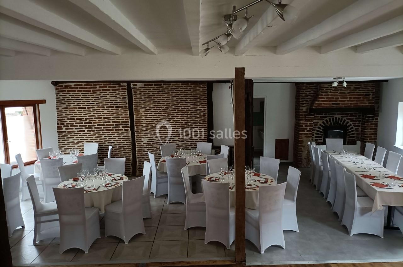 Salle de réception avec murs en briques, tables rondes et rectangulaires dressées pour un repas, chaises blanches.