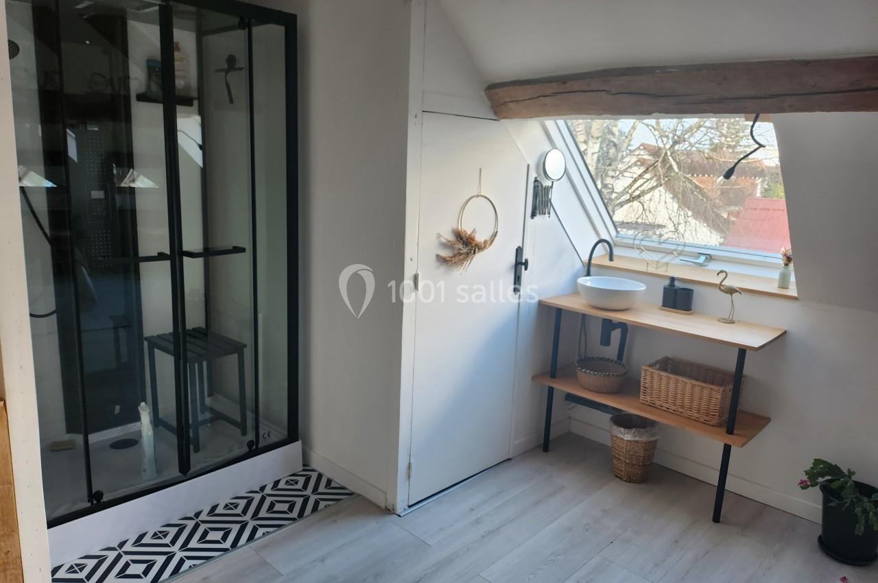Salle de bain lumineuse sous combles avec douche vitrée, lavabo sur meuble en bois et fenêtre donnant sur l'extérieur.