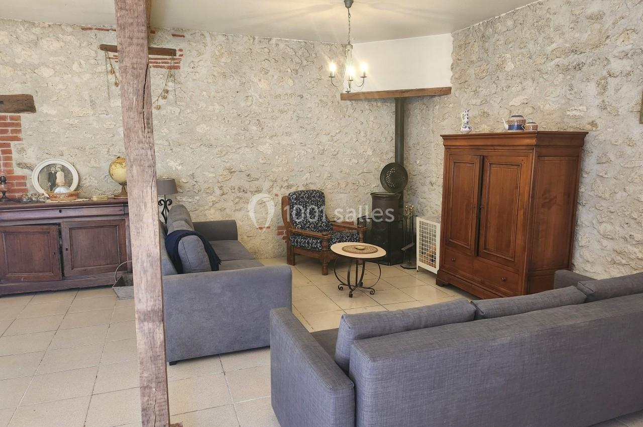 Salon avec murs en pierre, canapés gris, fauteuil, armoire en bois, table basse et éclairage suspendu.