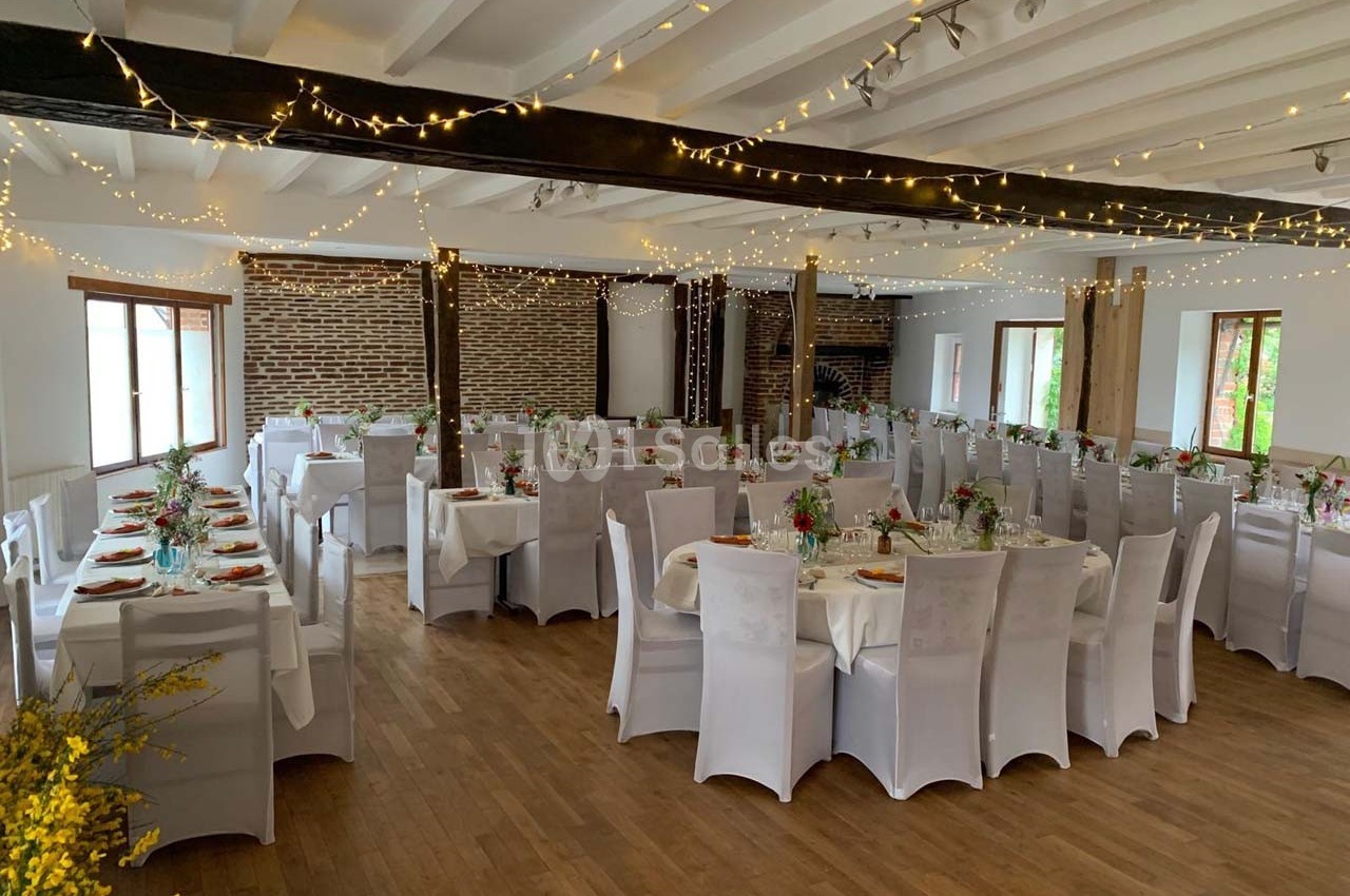 Salle de réception décorée avec des guirlandes lumineuses, tables dressées avec nappes blanches et chaises assorties.