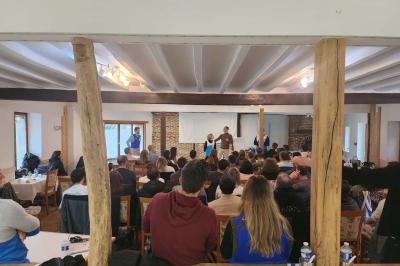 Un groupe de personnes assises et debout dans une salle avec des poutres apparentes, participant à une présentation.