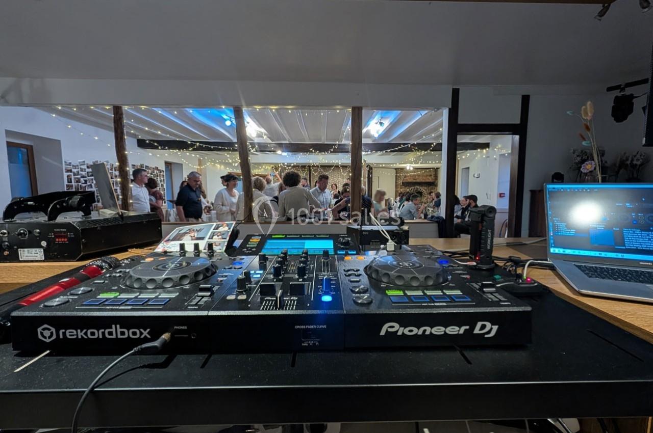 Vue d'une table de mixage DJ avec des invités en arrière-plan dans une salle décorée de guirlandes lumineuses.