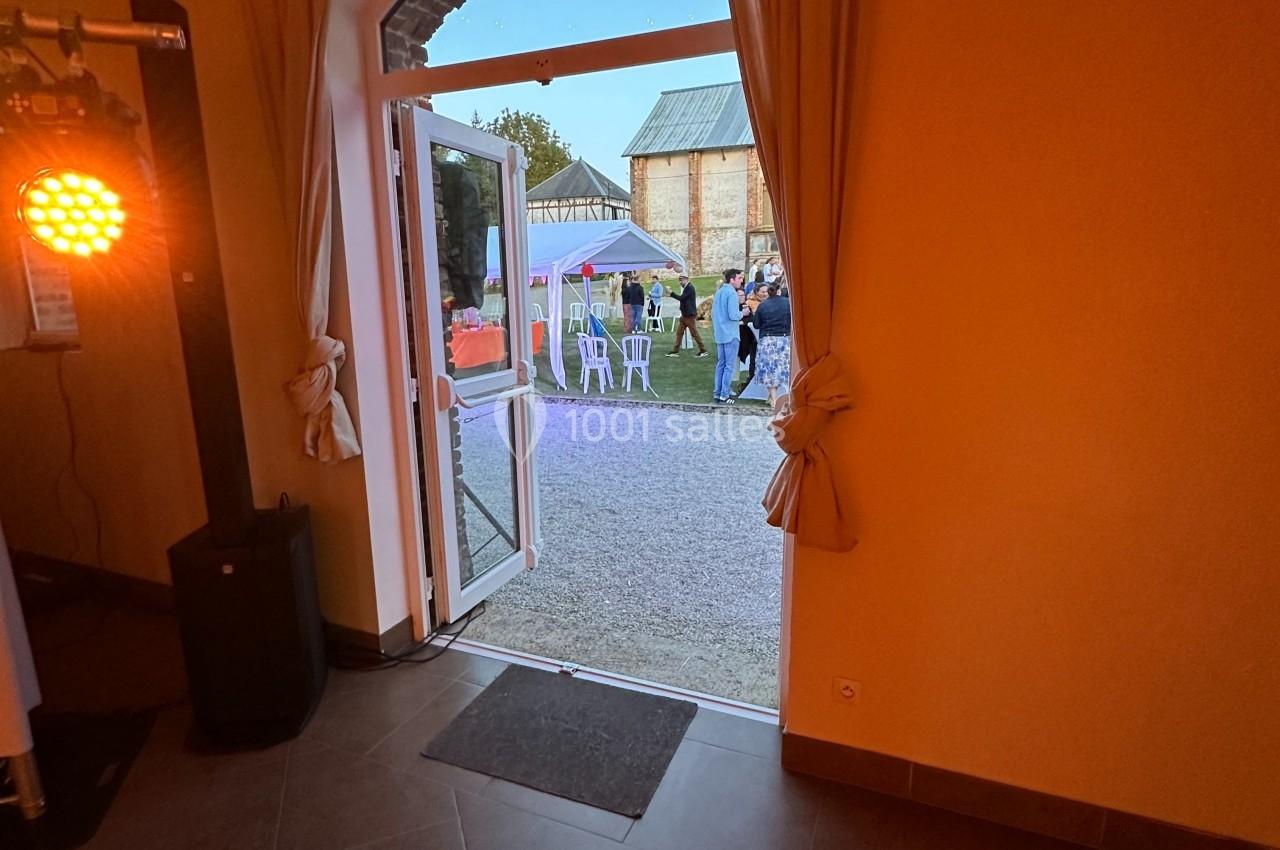 Vue depuis une salle éclairée vers une porte ouverte donnant sur un jardin avec des personnes et des tables en extérieur.