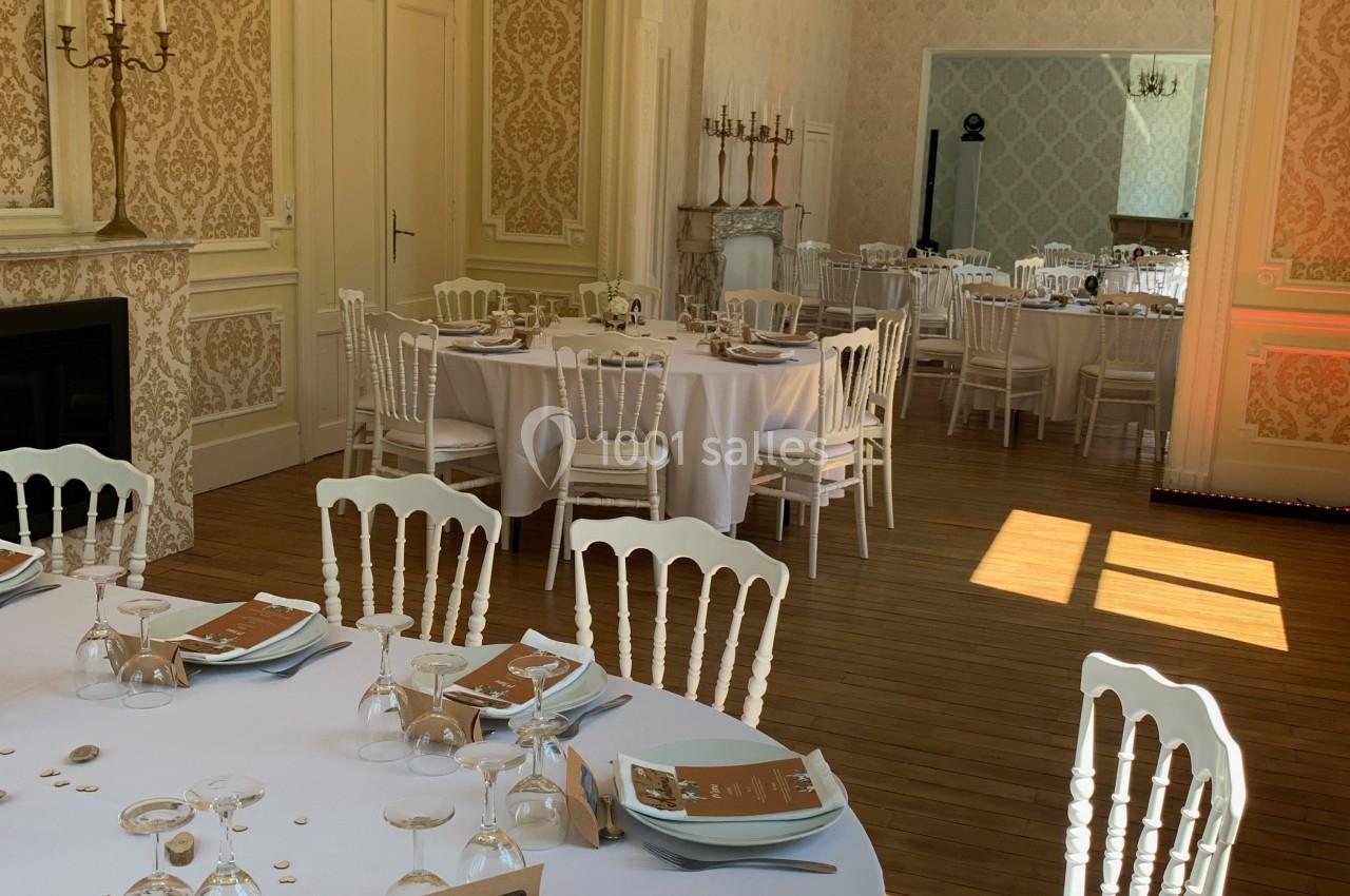 Salle élégante avec tables rondes dressées pour un événement, chaises blanches et lumière naturelle traversant les fenêtres.