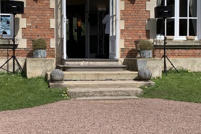 Entrée d'un bâtiment en briques avec porte ouverte, escaliers, pots de fleurs et haut-parleurs disposés de chaque côté.