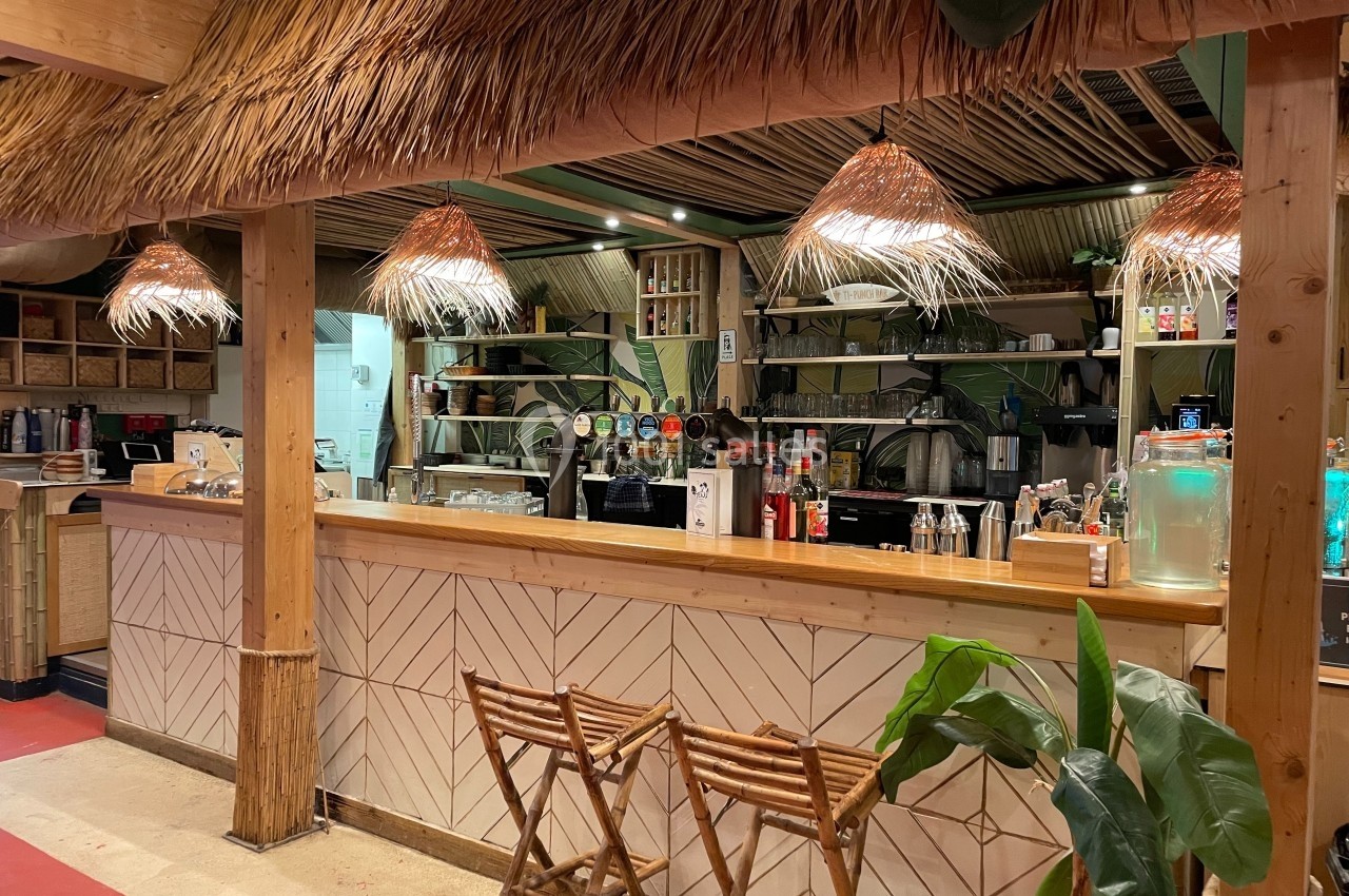 Comptoir en bois d'un bar à thème tropical avec tabourets pliants, éclairage en paille et décoration végétale.