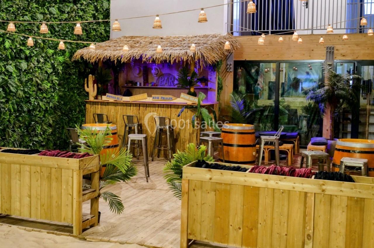 Bar en bois au style tropical avec toit en chaume, entouré de plantes, tonneaux et guirlandes lumineuses.
