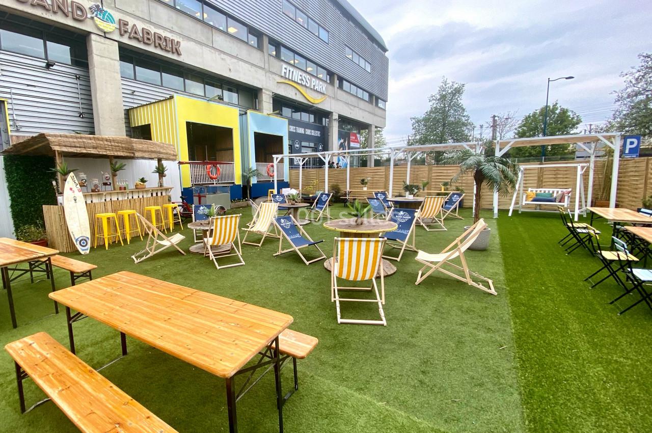 Espace extérieur aménagé avec chaises longues, tables en bois et décor tropical sur pelouse synthétique.