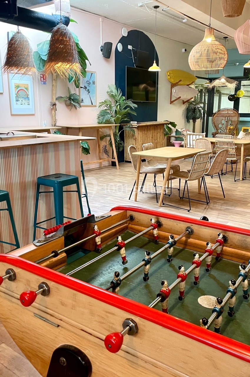 Baby-foot en bois dans un espace lumineux avec bar, tables en rotin, plantes et décorations suspendues.