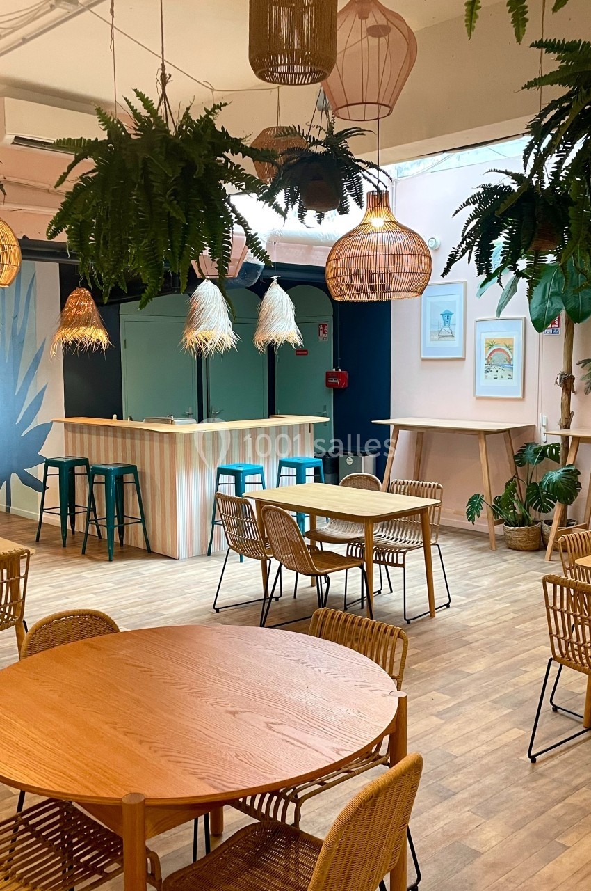 Salle lumineuse avec tables en bois, chaises en rotin, plantes suspendues et comptoir de bar en arrière-plan.
