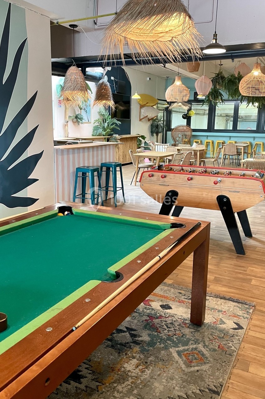 Salle conviviale avec une table de billard, un baby-foot, des tabourets et une décoration lumineuse et végétale.