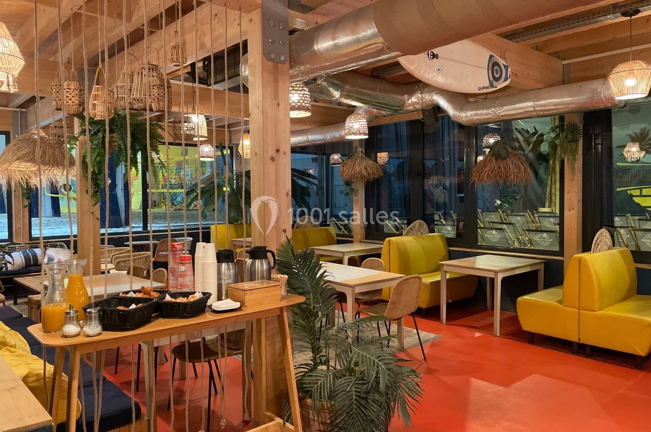 Salle de restaurant lumineuse avec tables en bois, banquettes jaunes, décor tropical et éléments suspendus en rotin.