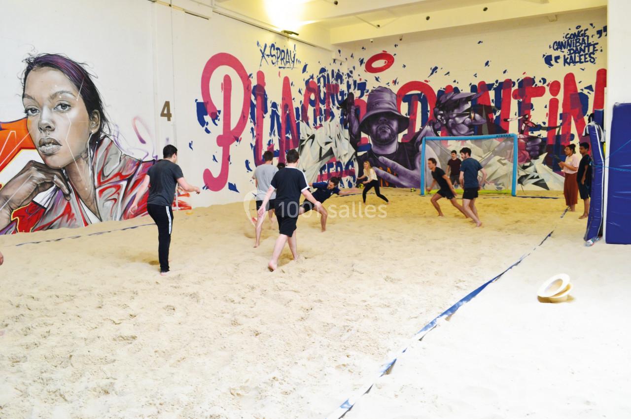 Des personnes jouent au football sur un terrain de sable intérieur, entouré de fresques murales colorées.