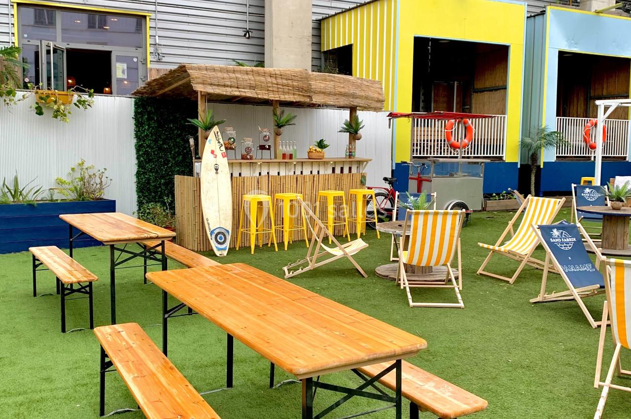 Tables en bois, chaises longues et bar en extérieur sur une terrasse avec gazon synthétique devant un bâtiment.