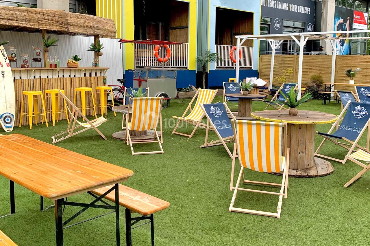 Terrasse extérieure avec chaises longues, tables en bois et décor tropical sur un sol en gazon synthétique.