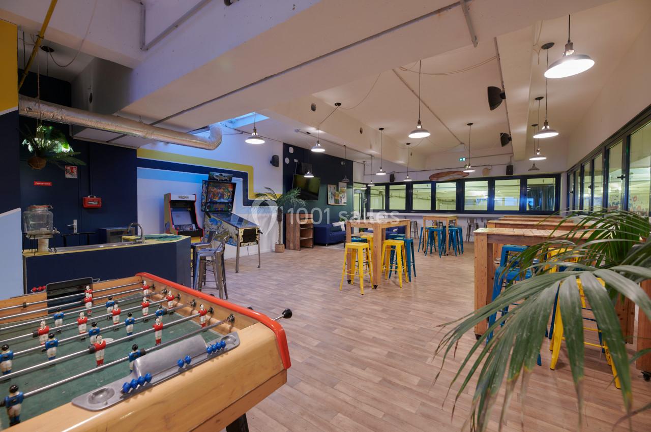 Salle de détente avec baby-foot, tables hautes, tabourets colorés et jeux d'arcade dans un espace lumineux.