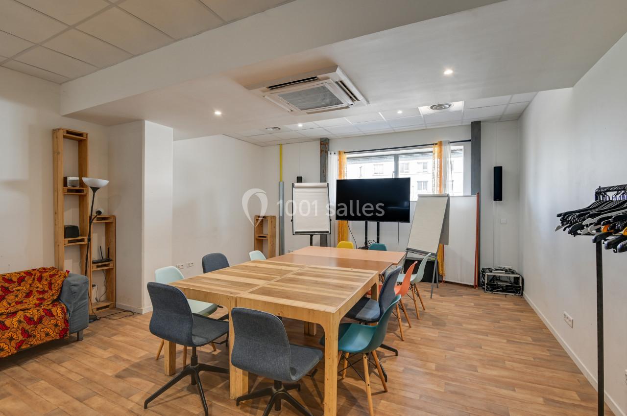 Salle de réunion lumineuse avec table en bois, chaises colorées, tableau blanc, écran et portants à vêtements.