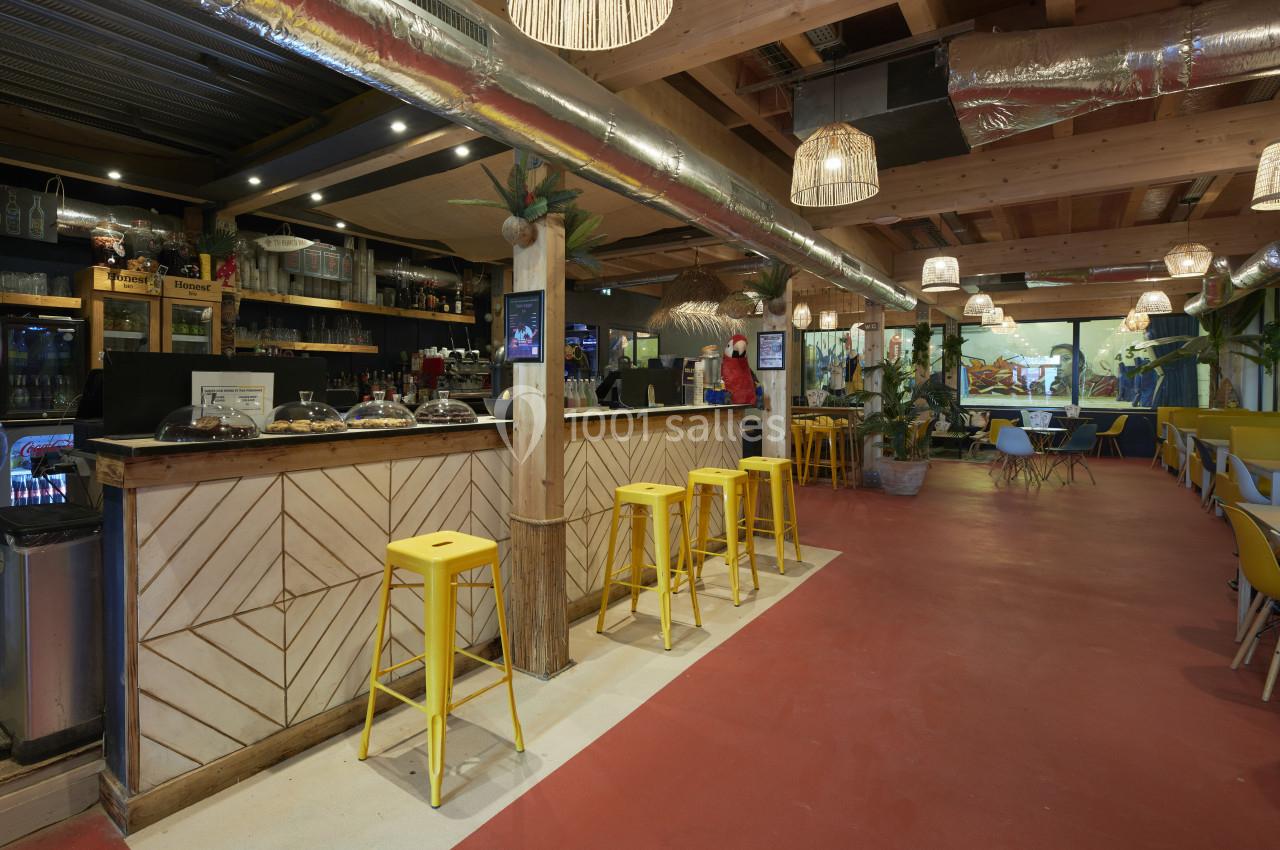 Intérieur d'un café avec un comptoir en bois, tabourets jaunes, éclairage suspendu et décoration végétale.