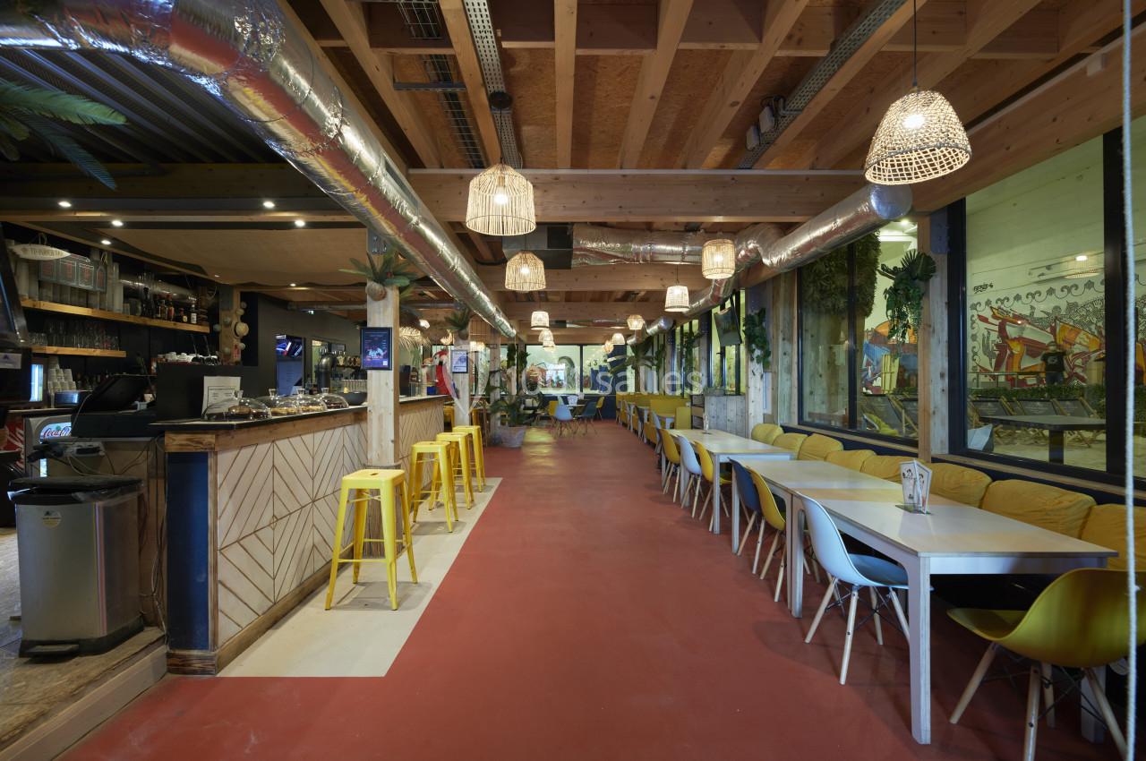 Intérieur d'un restaurant lumineux avec un bar en bois, des tables alignées et des chaises colorées.