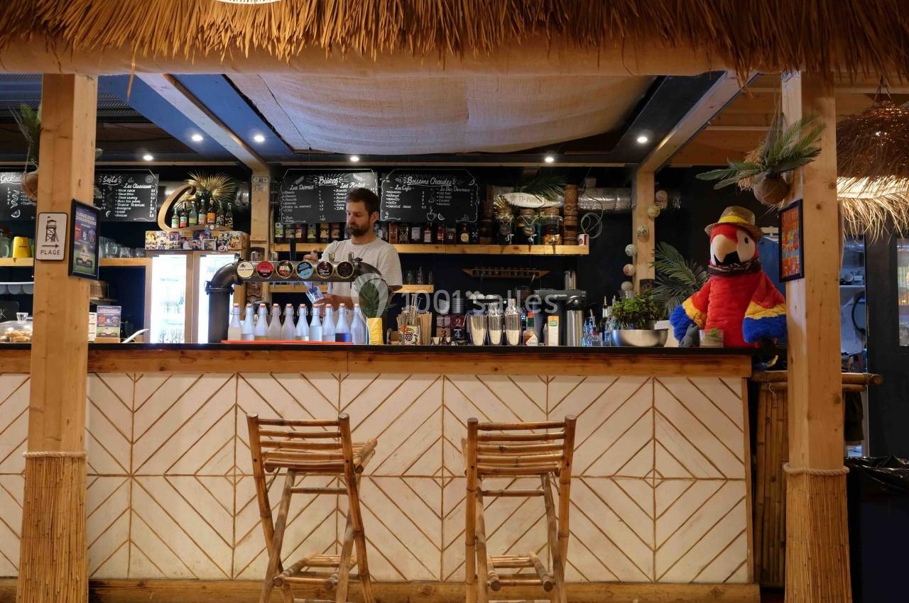 Bar tropical avec comptoir en bois, tabourets en bambou, décor exotique et barman servant des boissons.