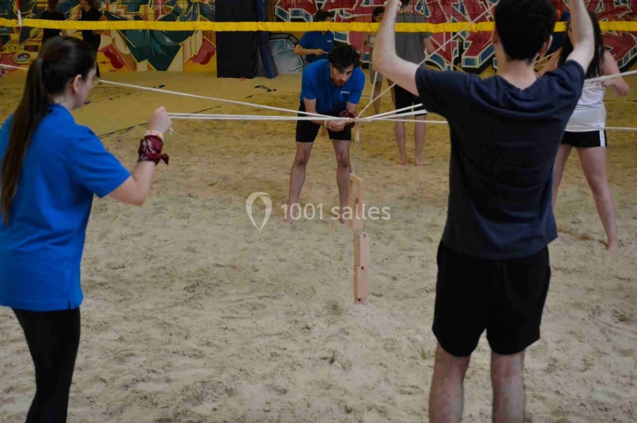 Des personnes collaborent pour empiler des blocs de bois à l'aide de cordes dans un espace intérieur sablonneux.