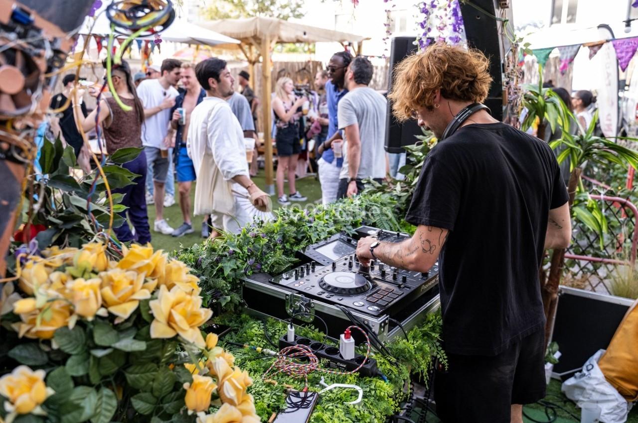 Un DJ mixe en plein air lors d'un événement festif, entouré de plantes et de participants discutant en arrière-plan.