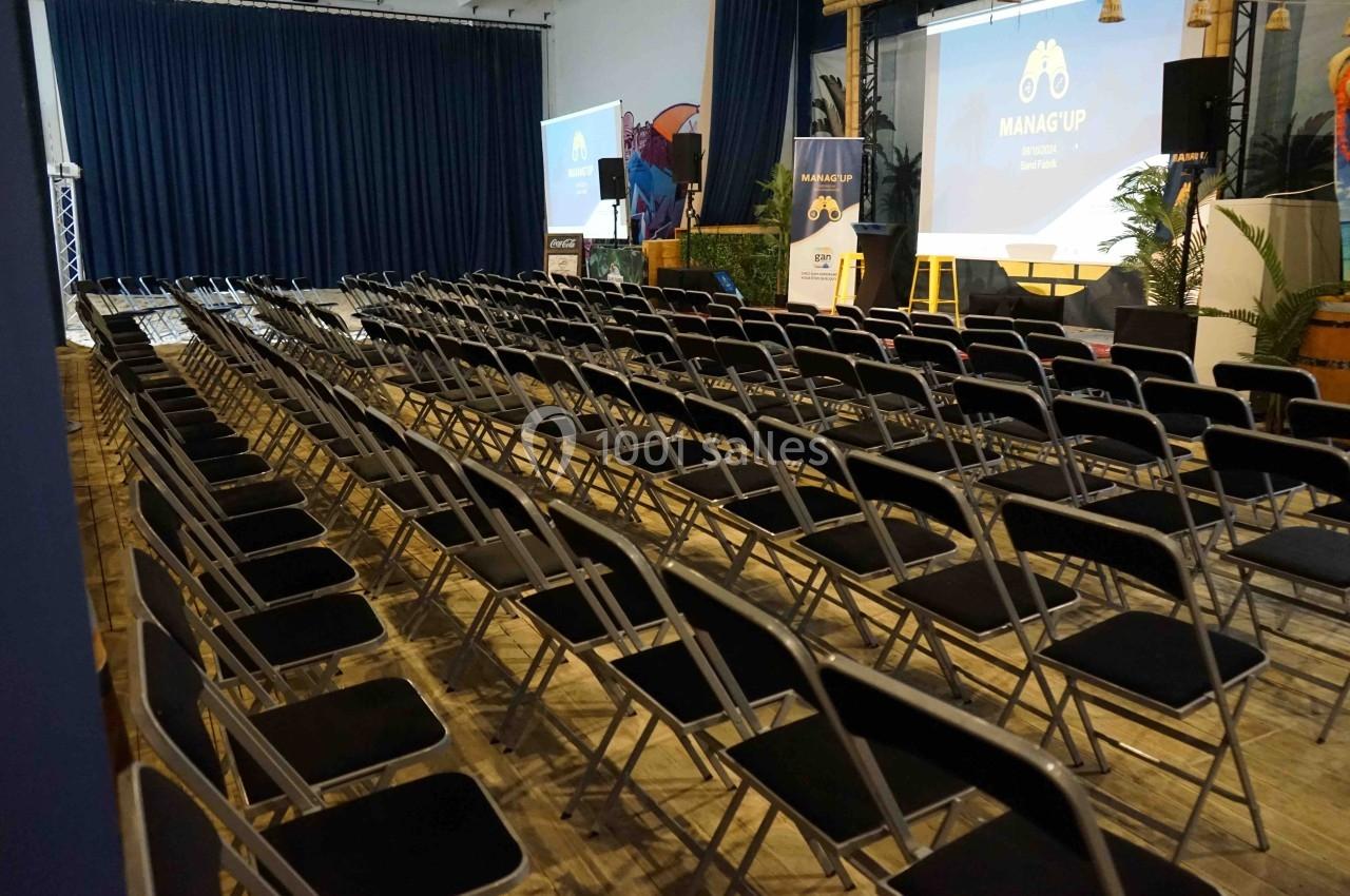 Salle de conférence avec des rangées de chaises noires vides, un écran de projection et des éléments de décoration.