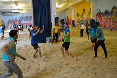 Des personnes participent à un jeu d'adresse avec des labyrinthes en bois dans un espace intérieur sur sol sablé.