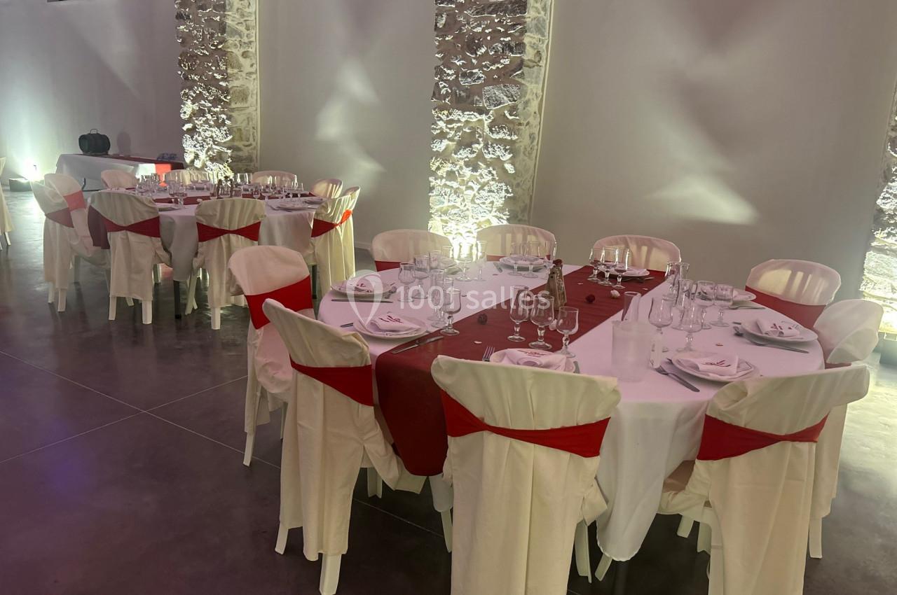 Tables dressées avec nappes blanches, chemins de table rouges et chaises décorées, dans une salle aux murs en pierre…
