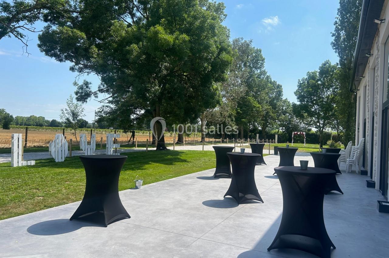 Terrasse extérieure avec tables hautes noires, vue sur un jardin arboré et un champ sous un ciel dégagé.