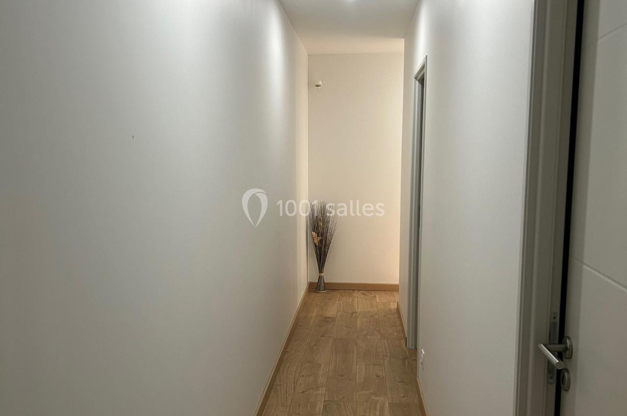 Couloir étroit avec murs blancs, sol en bois clair et vase décoratif avec branches au fond.