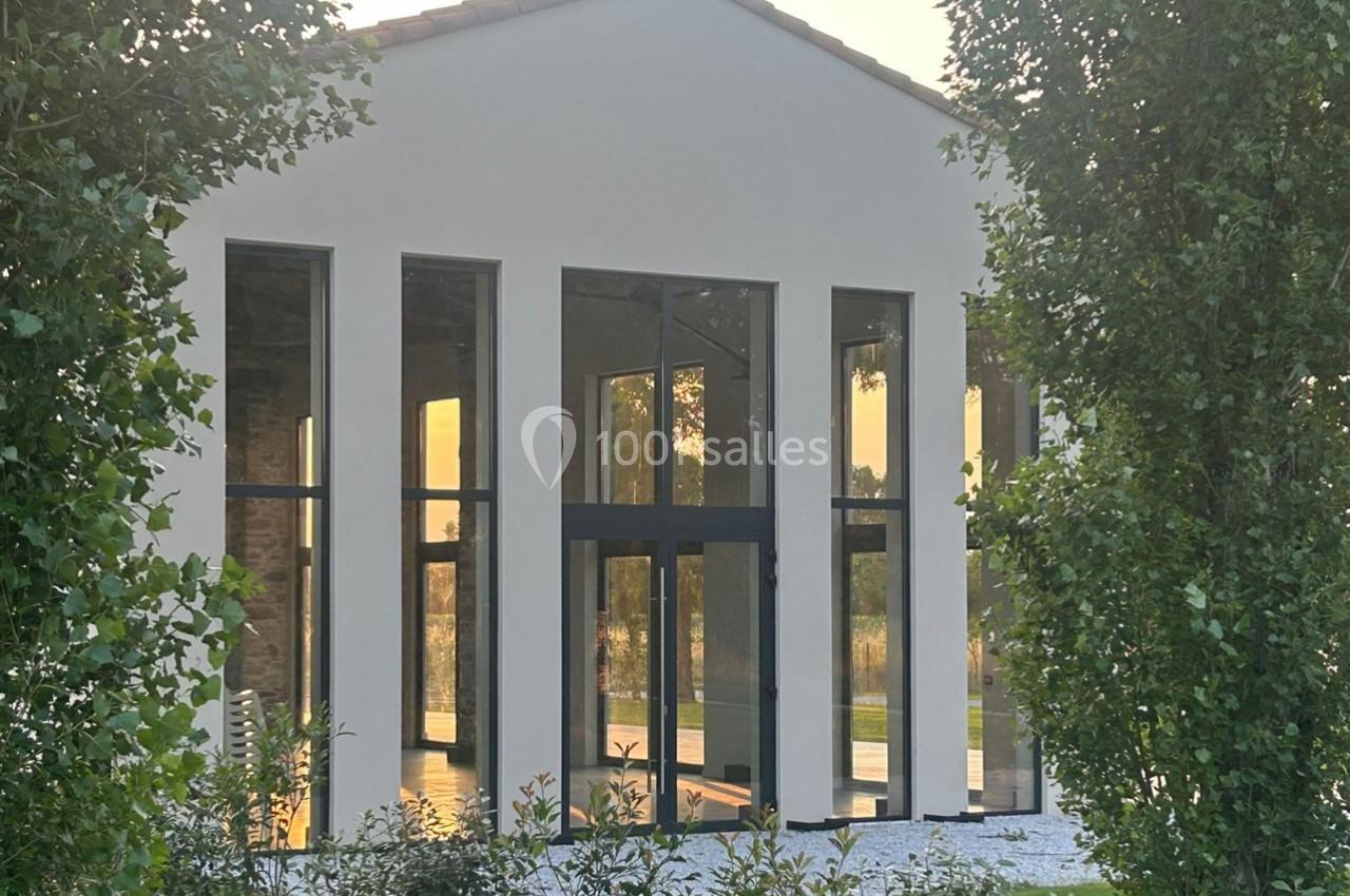 Façade d'une maison moderne avec de grandes baies vitrées, entourée de verdure et éclairée par une lumière douce.