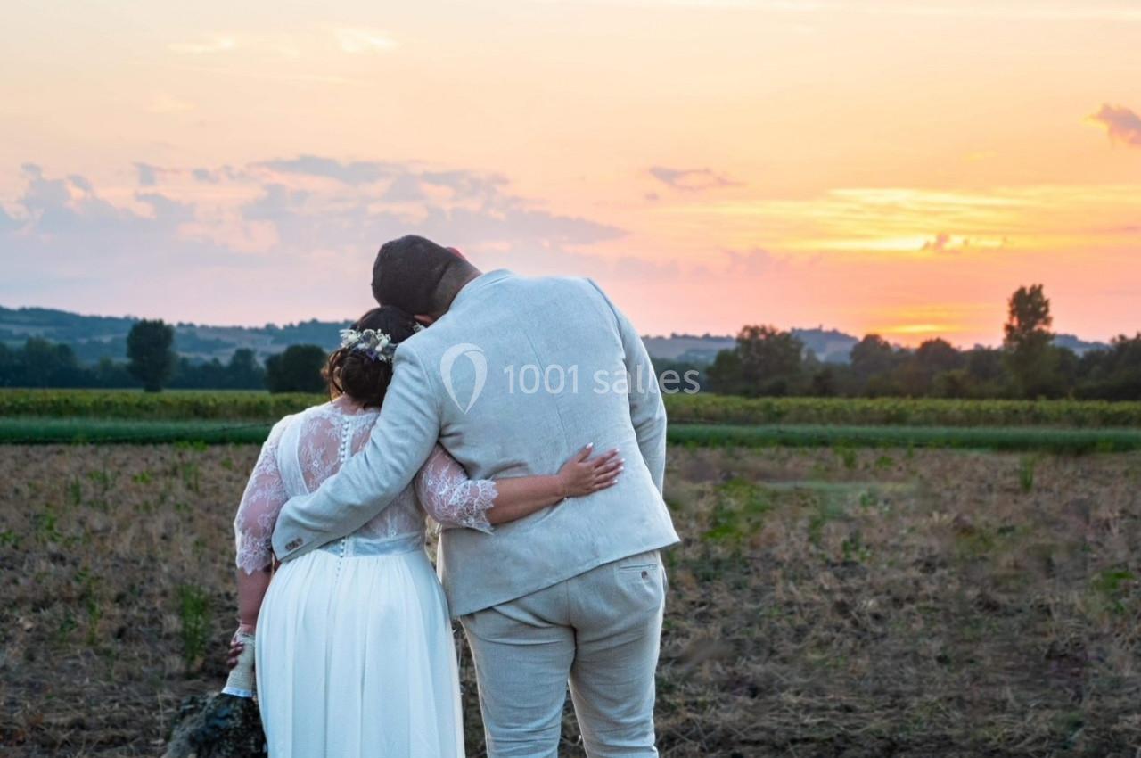 Un couple de dos, vêtu de tenues de mariage, regarde un coucher de soleil dans un paysage champêtre.