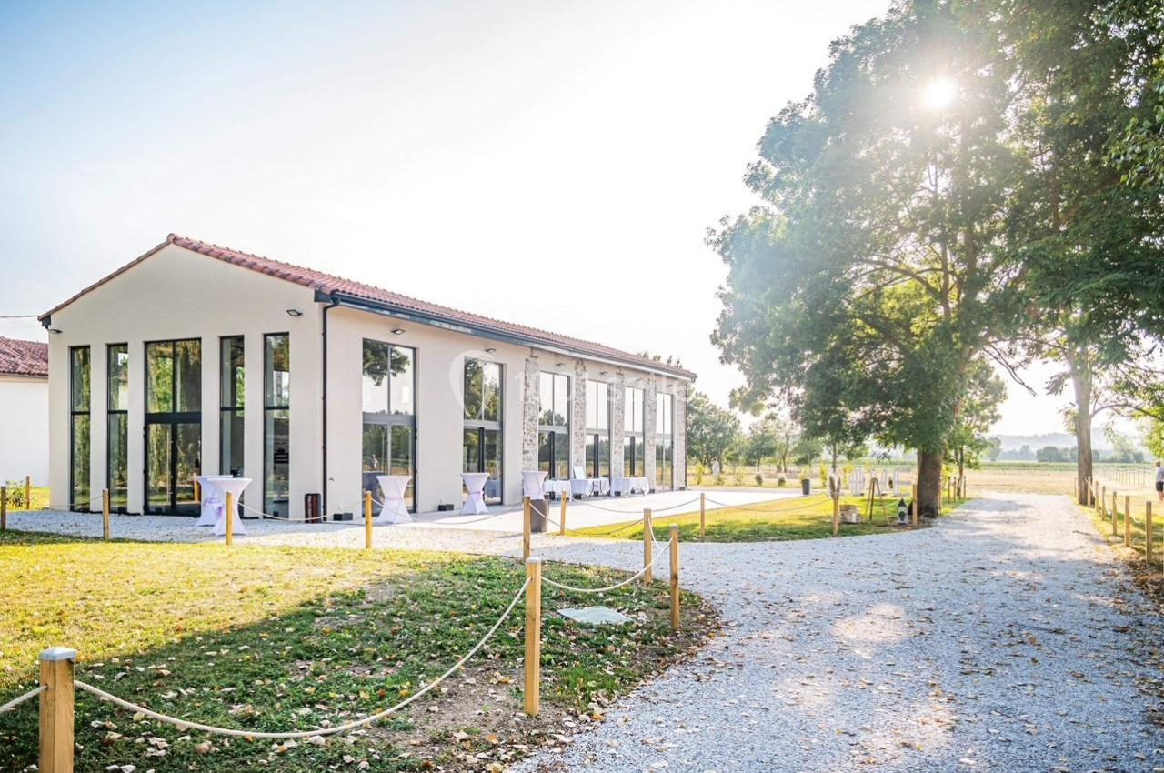 Bâtiment moderne avec de grandes baies vitrées, entouré d'arbres et d'un chemin gravillonné sous un ciel ensoleillé.