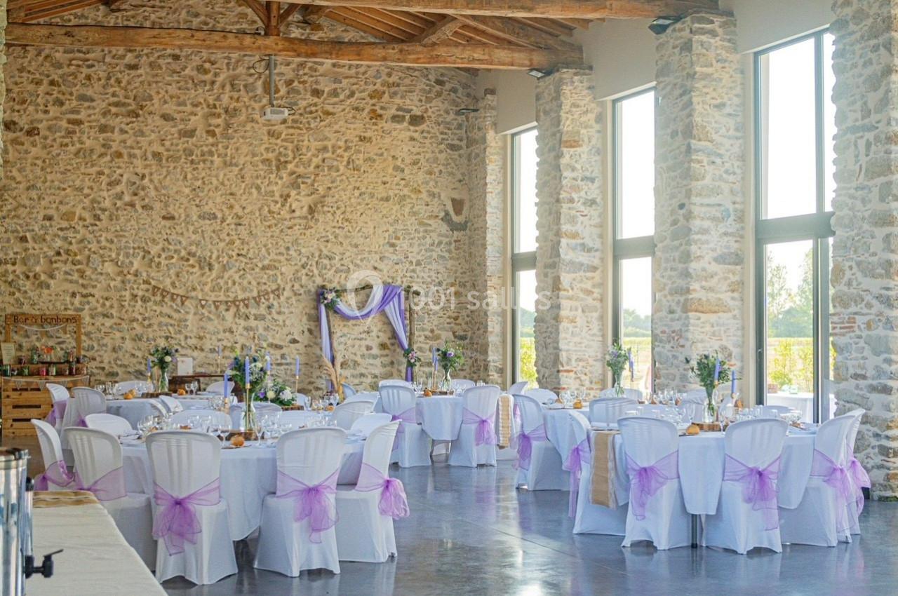 Salle de réception en pierre avec tables décorées de nappes blanches et chaises ornées de rubans violets.