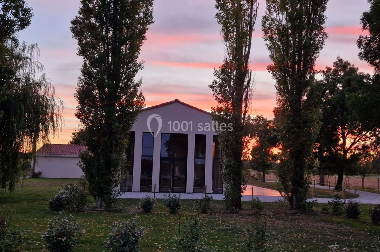 Façade d'une maison moderne entourée d'arbres, au coucher du soleil avec un ciel aux teintes roses et orangées.