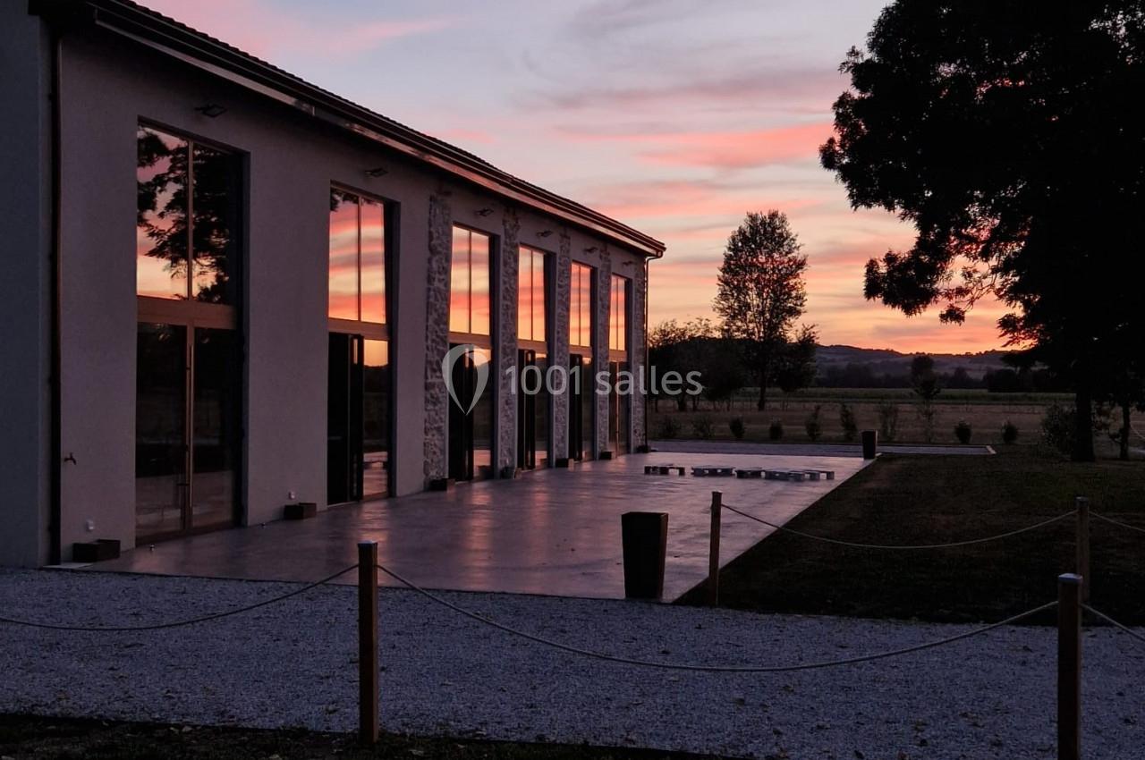 Bâtiment moderne avec de grandes baies vitrées reflétant un ciel rose au coucher du soleil, entouré d'arbres et d'un jardin.