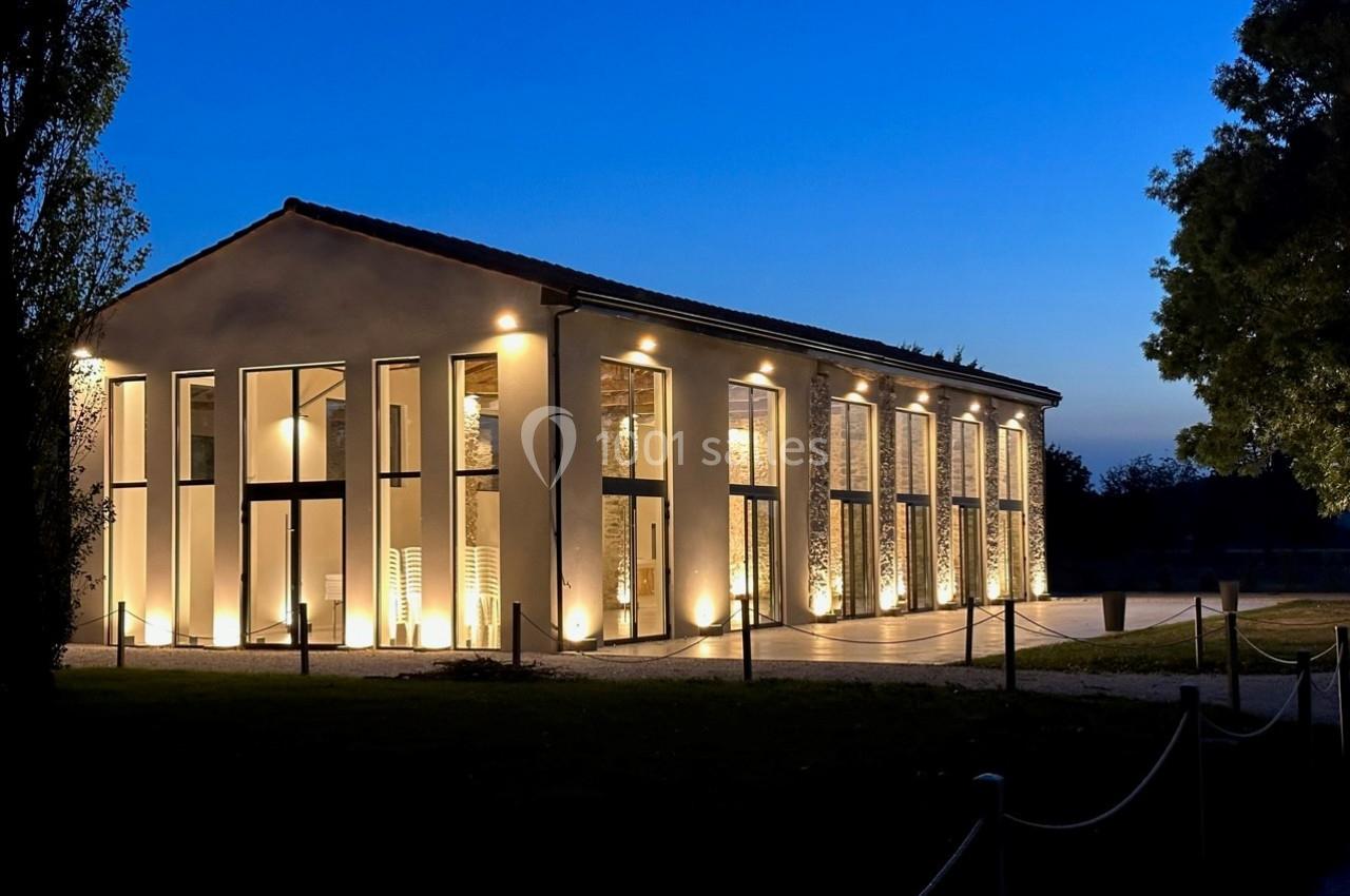 Bâtiment moderne éclairé de nuit, avec de grandes baies vitrées et entouré d'arbres.