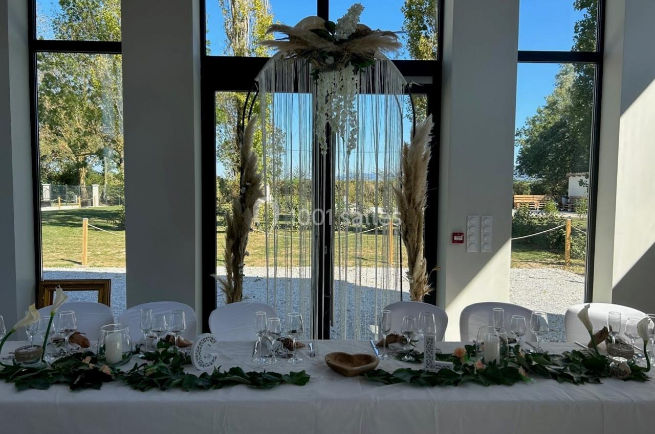 Table décorée pour un événement avec nappes blanches, vaisselle élégante, feuillage et arche ornée de pampas en arrière-plan.