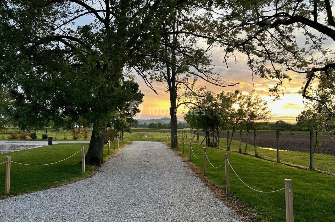 Allée de gravier bordée d'arbres et de pelouses, menant à un paysage de campagne au coucher du soleil.