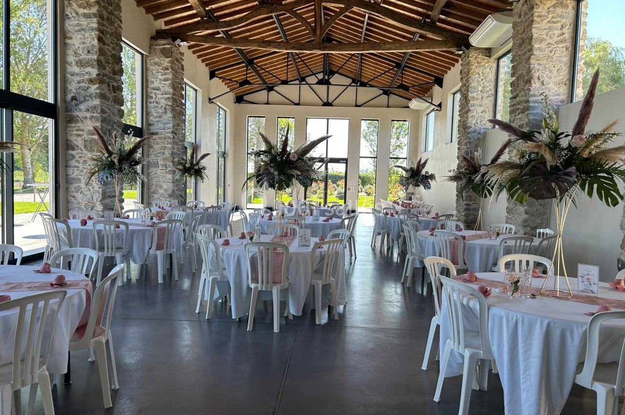 Salle de réception lumineuse avec tables rondes dressées, décorations florales et poutres apparentes au plafond.