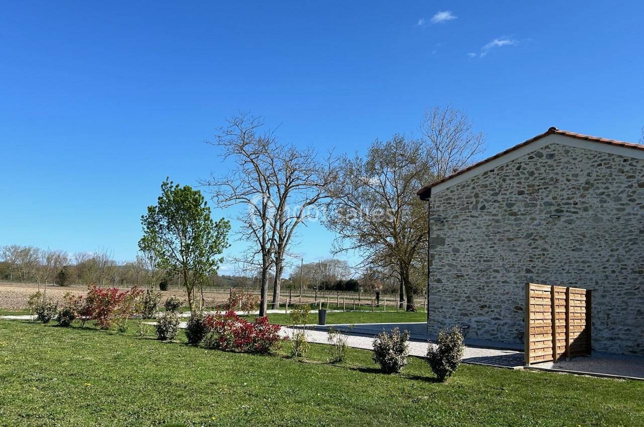 Jardin avec pelouse, arbustes et arbres, bordé par un bâtiment en pierre sous un ciel bleu dégagé.
