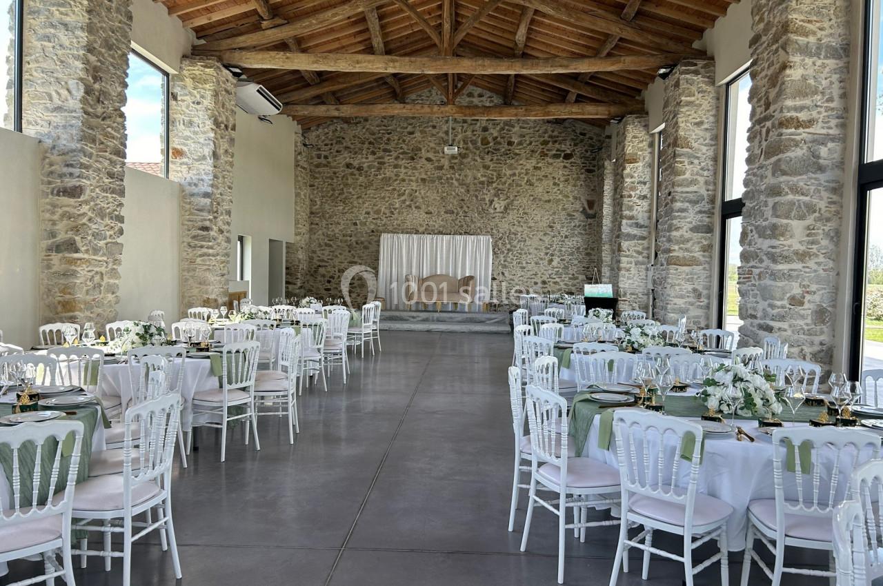 Salle de réception en pierre avec tables rondes dressées, chaises blanches et une petite scène en fond.