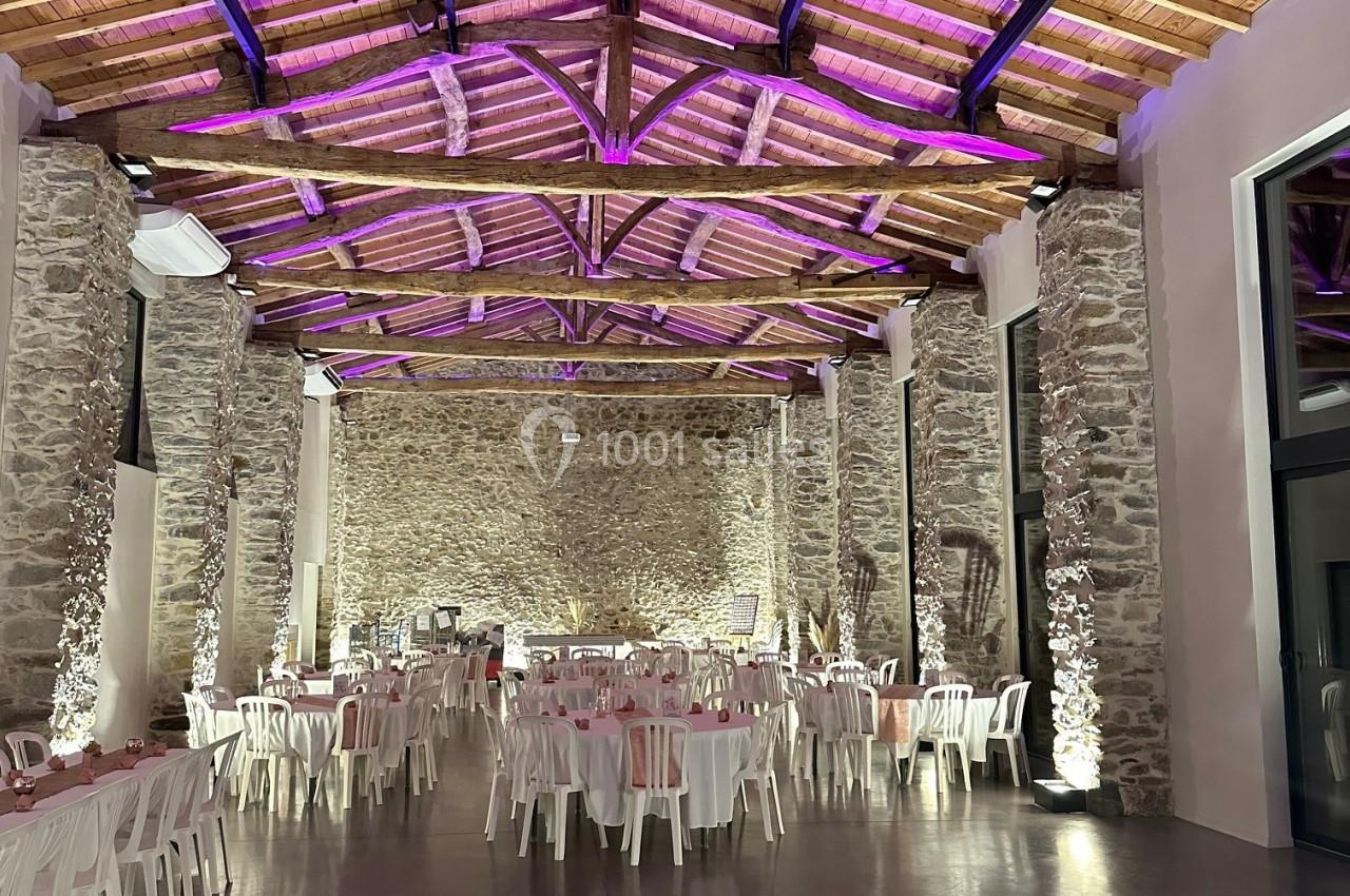 Grande salle en pierre avec plafond en bois, éclairage violet et tables blanches disposées pour un événement.