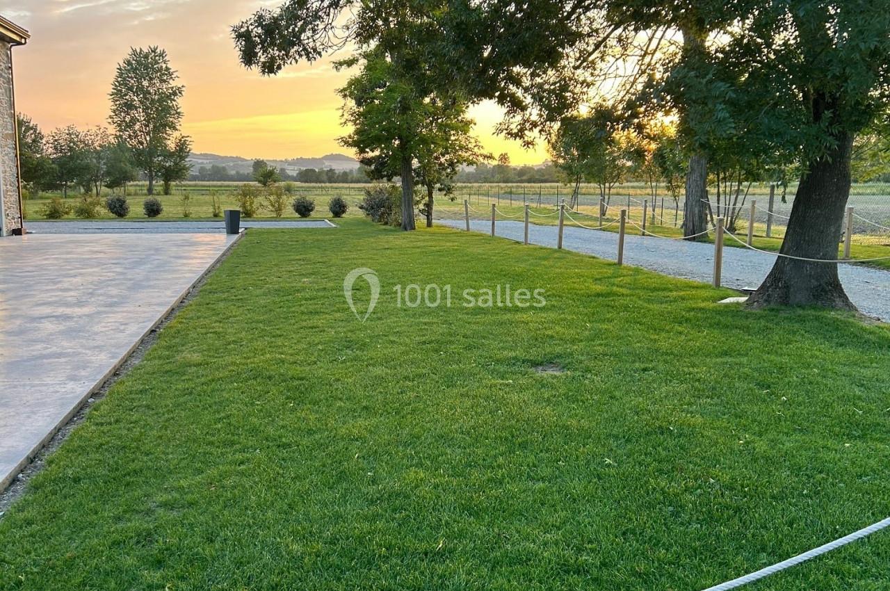 Pelouse verte bordée d'arbres au coucher du soleil, avec une terrasse à gauche et une clôture en arrière-plan.
