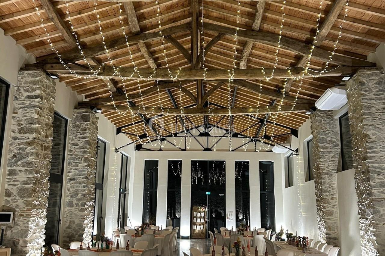 Salle de réception avec des tables décorées, des chaises blanches et un plafond en bois orné de guirlandes lumineuses.