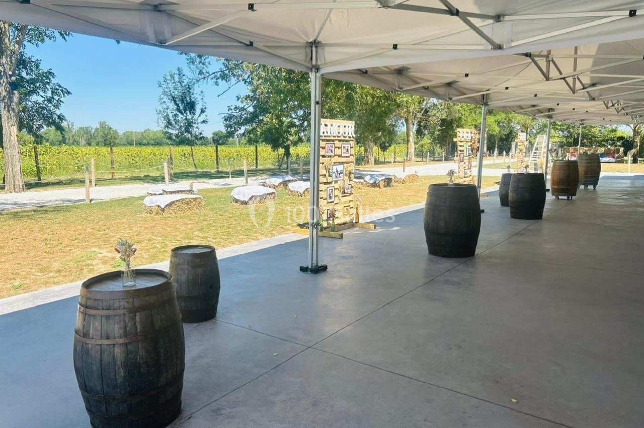 Tonelle blanche installée en extérieur avec des tonneaux en bois servant de tables, entourée de verdure.